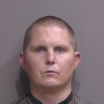 A man with short light brown hair and fair skin looks directly at the camera with a neutral expression, standing against a plain gray background