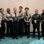 A group of ten uniformed officers stands together indoors Two in the center hold a plaque and a large trophy The group is smiling, posing in front of a plain wall with a blue patterned carpet