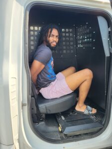 A man with dreadlocks, wearing a graphic t shirt, pink shorts, and slide sandals, sits with his hands behind his back in the back seat of a police van with a partition behind him