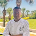 A chef in a white uniform stands outside with arms crossed, palm trees and greenery in the background on a sunny day