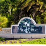A stone and tile welcome sign for Palm Coast featuring white text and a palm tree graphic, surrounded by green foliage and palm trees in the background
