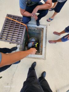 Several people stand around an open manhole, looking down at a person wearing a helmet and protective gear who is descending inside One person points towards the individual in the manhole