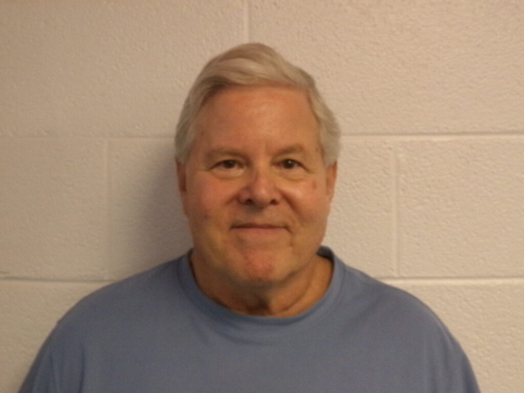 An older man with short gray hair smiles softly while standing against a light colored, textured brick wall He is wearing a light blue shirt