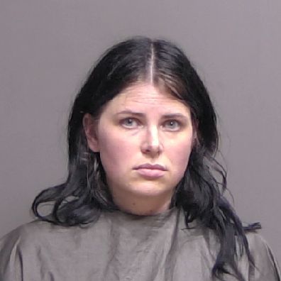 A woman with long dark hair and a serious expression is wearing a draped garment, posed against a plain, neutral colored background for a mugshot style photo