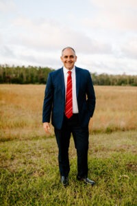 A man in a blue suit, white shirt, and red striped tie stands smiling in a grassy field with trees and a cloudy sky in the background