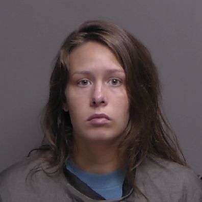A woman with long brown hair and a serious expression is pictured against a plain, dark background, wearing a dark colored garment draped around her shoulders