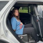 A person with long brown hair, wearing a blue T shirt and black pants, sits handcuffed in the back seat of a police vehicle, looking toward the camera with a neutral expression