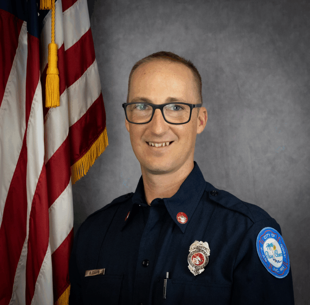 A firefighter in a navy blue uniform with badges and a patch stands in front of a U S flag and a gray backdrop, smiling at the camera and wearing glasses
