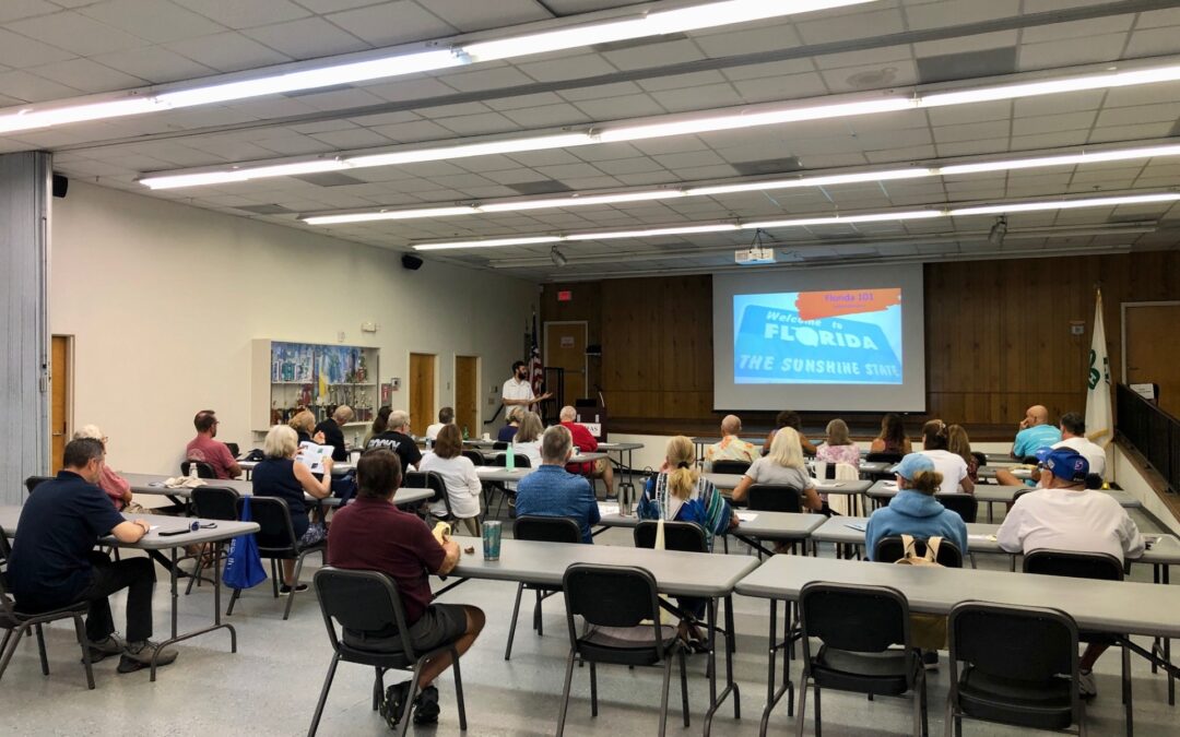 A group of people sit at tables in a large room, facing a presenter and a projection screen that reads "Welcome to Florida: The Sunshine State " The room has fluorescent lights and wood paneled walls