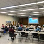 A group of people sit at tables in a large room, facing a presenter and a projection screen that reads "Welcome to Florida: The Sunshine State " The room has fluorescent lights and wood paneled walls