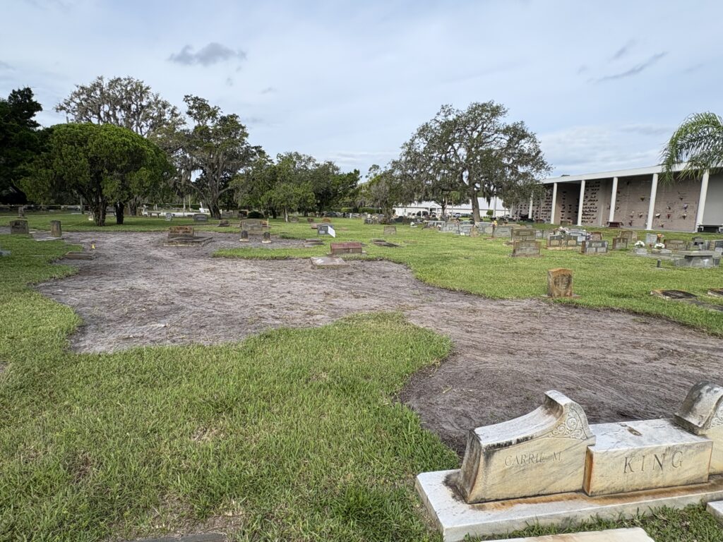 A cemetery with numerous headstones on green grass in Flagler A large, uneven patch of bare soil is in the foreground, with several damaged or fallen gravestones Trees and a white building are seen in the background under a cloudy sky
