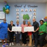 Seven people stand indoors, smiling and holding two oversized checks They are in front of a "Board of Directors" wall display and colorful balloons The setting appears to be an office or community organization