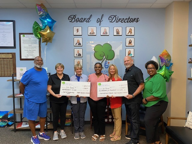 Seven people stand indoors, smiling and holding two oversized checks They are in front of a "Board of Directors" wall display and colorful balloons The setting appears to be an office or community organization