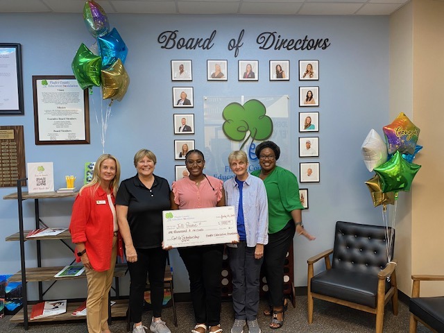 Five women stand smiling and holding a large check in front of a “Board of Directors” photo wall Colorful balloons are on both sides, and a green tree logo is on the wall behind them