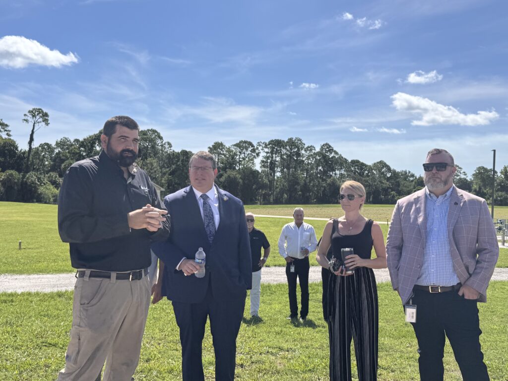 Four adults stand outdoors on grass under a blue sky, listening to a man in a black shirt speaking Trees line the background, and several other people stand farther behind the group