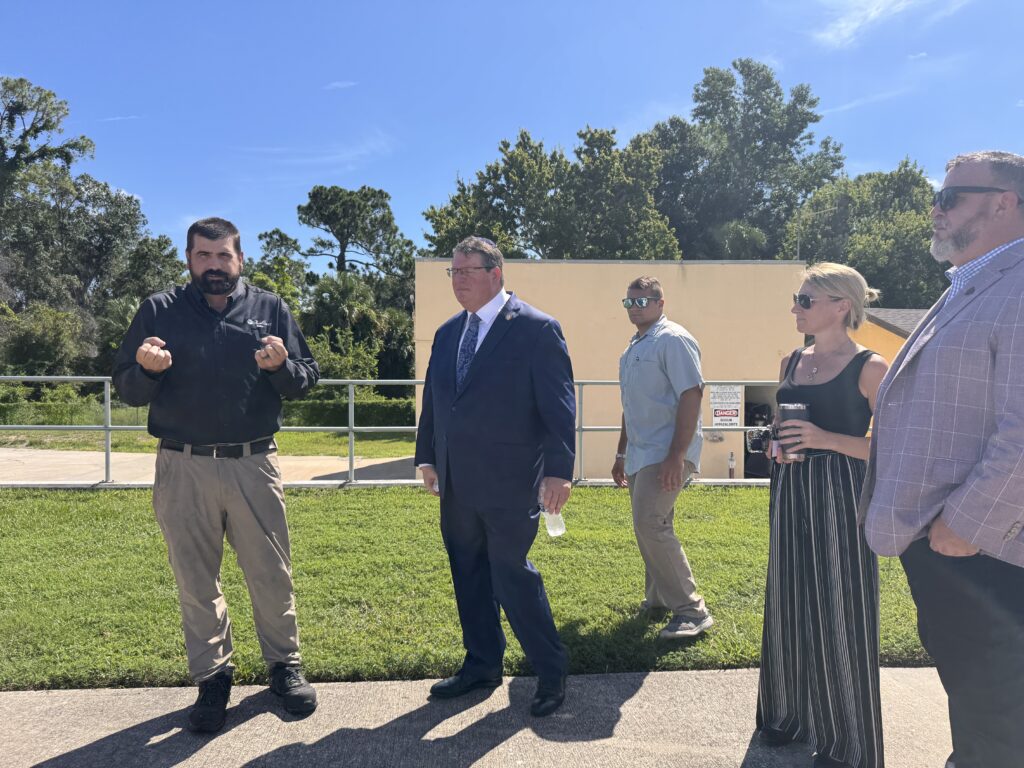 A group of five adults, including one man in a suit, stand outside on a sunny day; trees and a building are in the background One man gestures with his hands while talking to the group