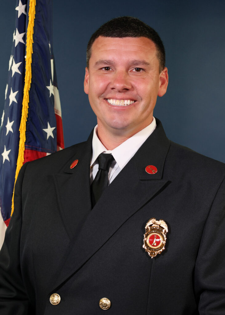 A smiling man in a formal dark firefighter uniform stands in front of a U S flag, wearing a badge and black tie, with a blue background behind him