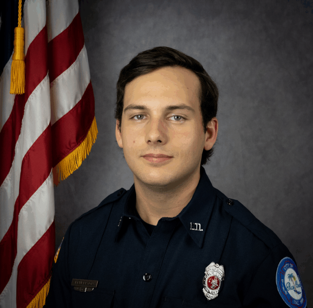 A young man in a dark blue uniform poses for a portrait in front of a U S flag and a gray backdrop He has short brown hair and wears a badge and a nameplate, indicating he is a public safety officer