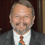 A middle aged man with a beard and mustache is smiling, wearing a pinstripe suit, white shirt, and a colorful tie The background is dark and out of focus