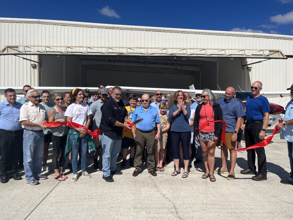 A group of people gathered outside a hangar, smiling and applauding as a ribbon is being cut in front of a small airplane, celebrating an event on a sunny day