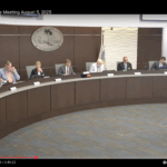 Seven Palm Coast city council members sit at a curved desk during a business meeting Papers and microphones are in front of them, and an American flag is visible A digital time display is on the desk