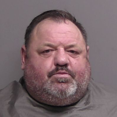 A middle aged man with short dark hair and a gray beard is facing the camera against a plain background, wearing a gray drape around his shoulders