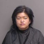 A woman with medium length black hair and a neutral expression wears a black cape against a plain gray background, as seen in a mugshot style photo