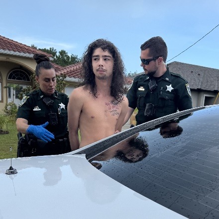 A shirtless man with long hair and visible chest tattoos is handcuffed and standing between two police officers in uniform next to a white car, outside a house on a cloudy day