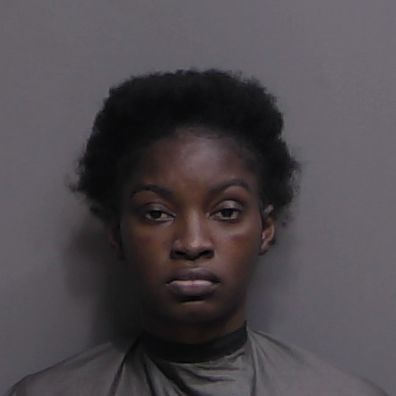 A woman with short, natural black hair and a serious expression faces the camera against a plain gray background, wearing a dark drape or garment A woman with short, natural black hair and a serious expression faces the camera against a plain gray background, wearing a dark drape or garment