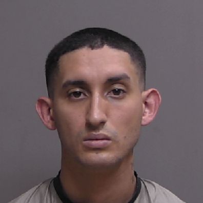 A man with short dark hair wearing a beige top is photographed against a plain gray background in a mugshot style image