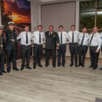 A group of twelve firefighters and officials, dressed in uniforms, stand in a line and smile for a group photo indoors, with a sunset painting and large windows in the background