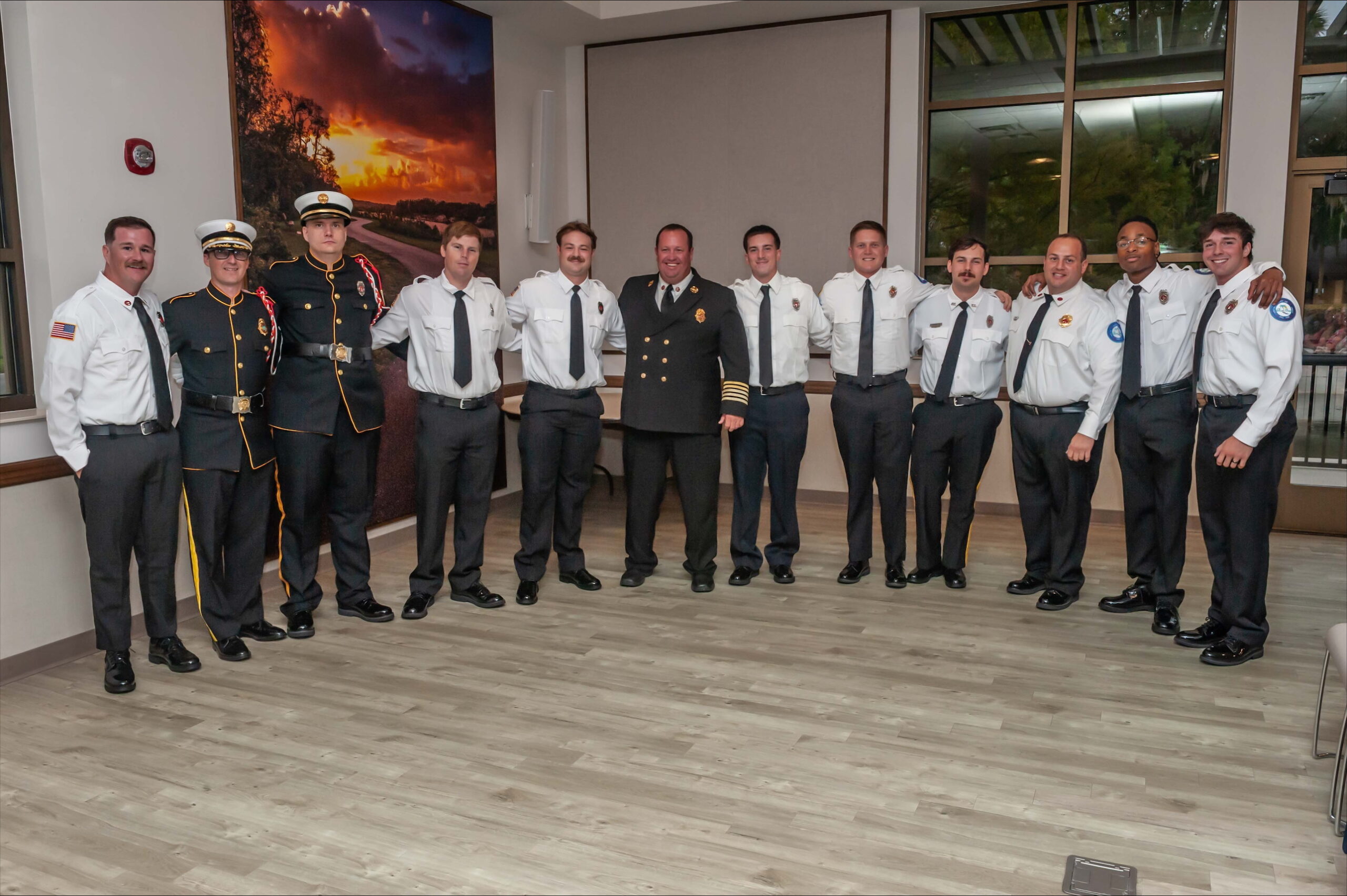 A group of twelve firefighters and officials, dressed in uniforms, stand in a line and smile for a group photo indoors, with a sunset painting and large windows in the background