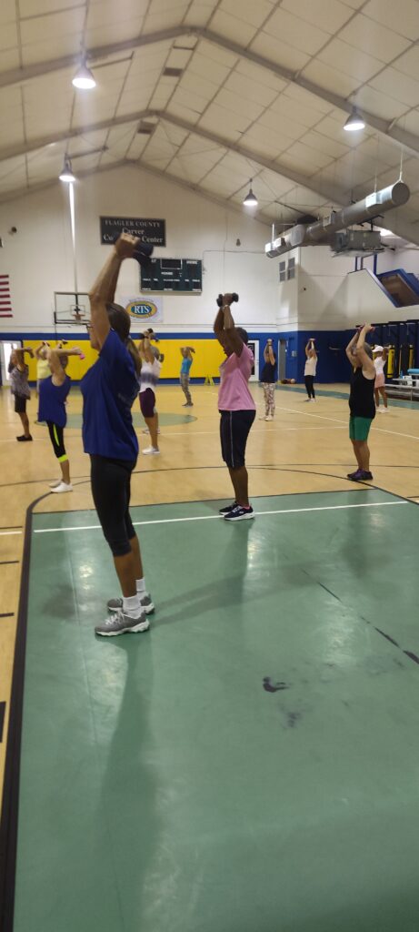 A group of people in athletic wear exercise together in a gymnasium, standing on basketball courts with arms raised, appearing to follow a fitness class