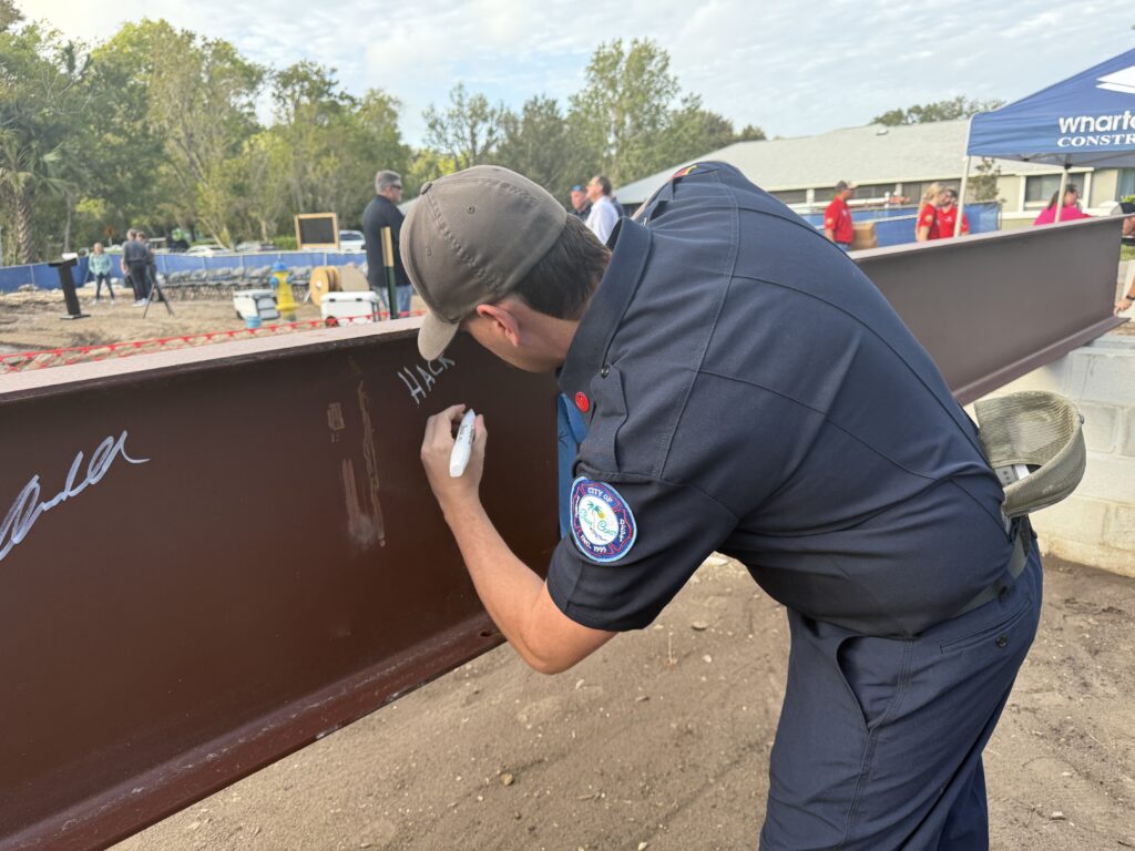 A person in a navy uniform and cap writes with a white marker on a large steel beam at an outdoor construction site, while others stand in the background near a blue tent