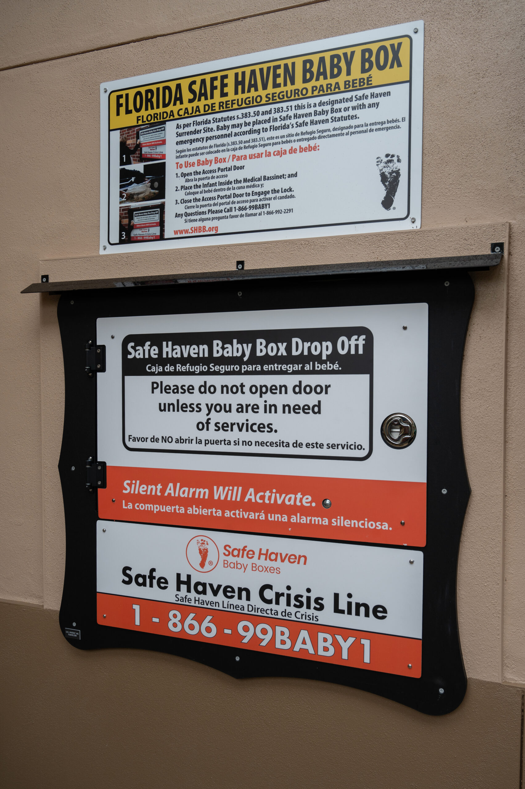 A Safe Haven Baby Box is installed on a wall with multiple signs in English and Spanish, providing a hotline number and instructions for safely surrendering a baby