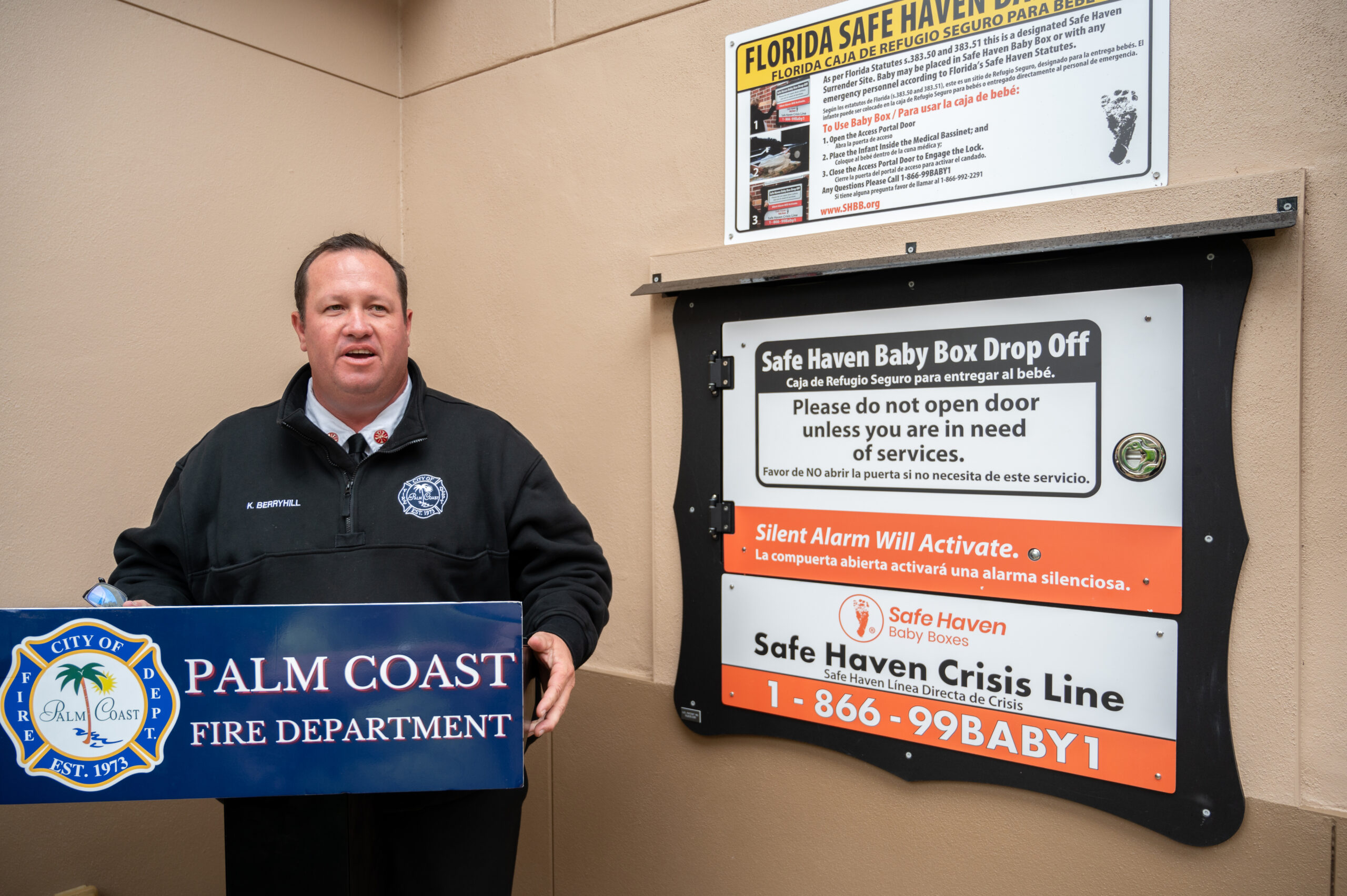 A man in a Palm Coast Fire Department jacket speaks at a podium beside Safe Haven Baby Box Drop Off signs and a Florida Safe Haven law notice, promoting baby safe surrender options