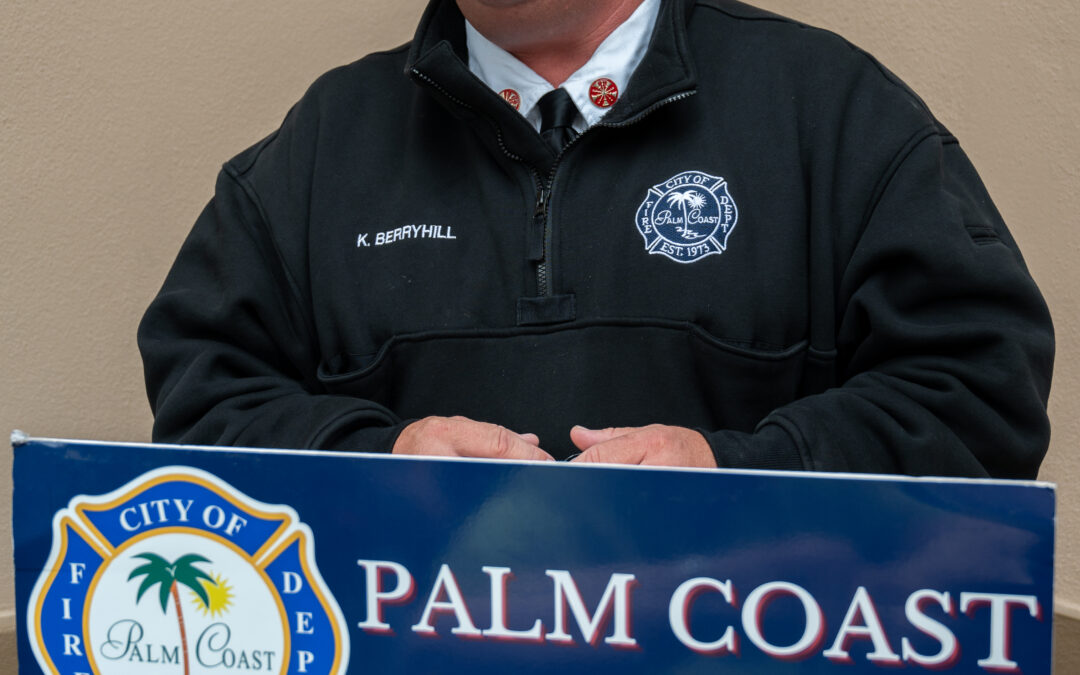 A man in a black fire department jacket stands at a podium that reads "Palm Coast Fire Department " He is speaking and smiling, with a beige wall in the background