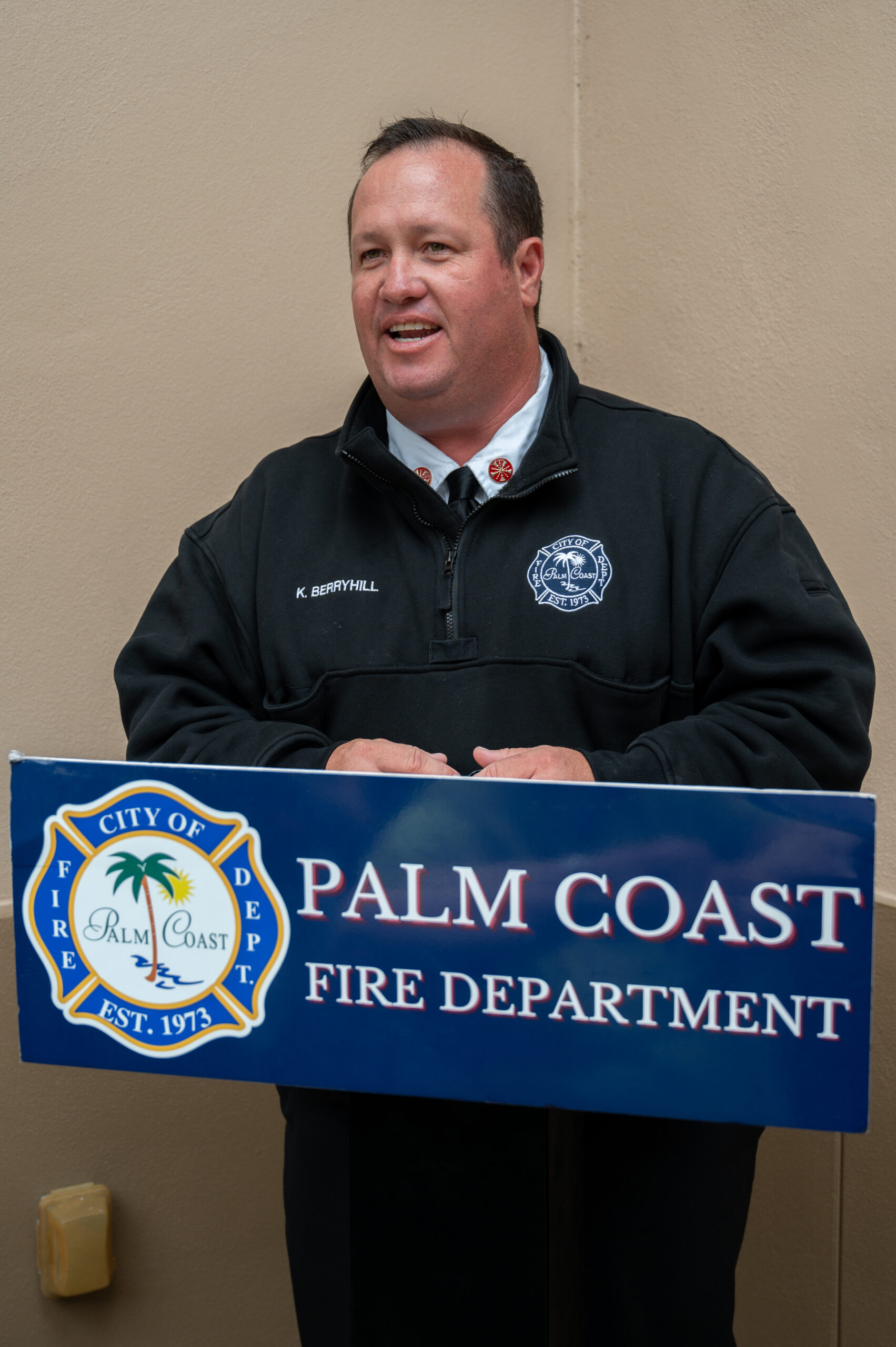 A man in a black fire department jacket stands at a podium that reads "Palm Coast Fire Department " He is speaking and smiling, with a beige wall in the background