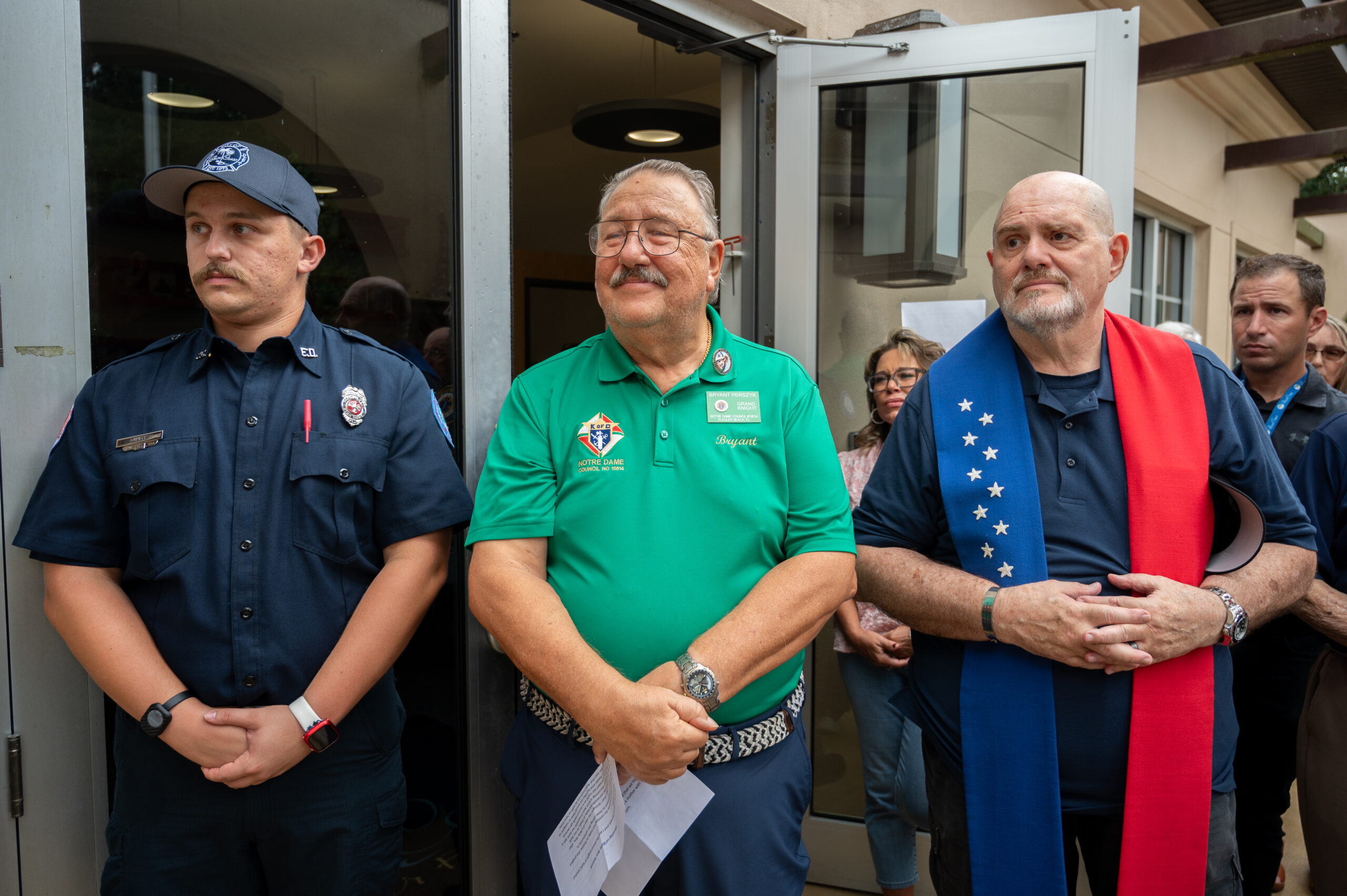 Three men stand side by side at an event; one wears a firefighter uniform, one wears a green polo with badges and documents, and one wears a dark shirt with a red, white, and blue scarf People are gathered behind them