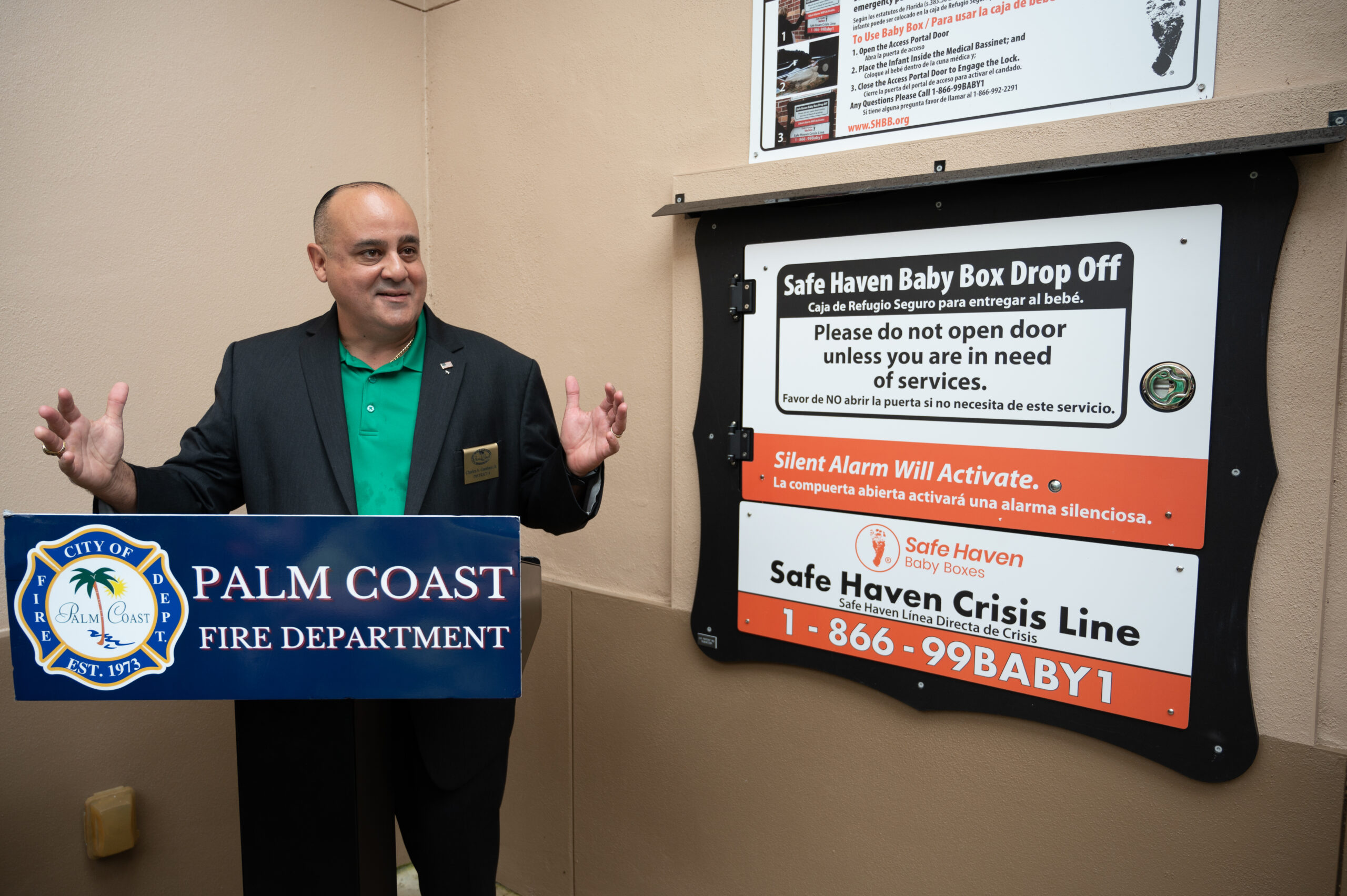 A man in a suit speaks at a podium labeled "Palm Coast Fire Department" next to a Safe Haven Baby Box Drop Off with instructions and crisis line information on the wall