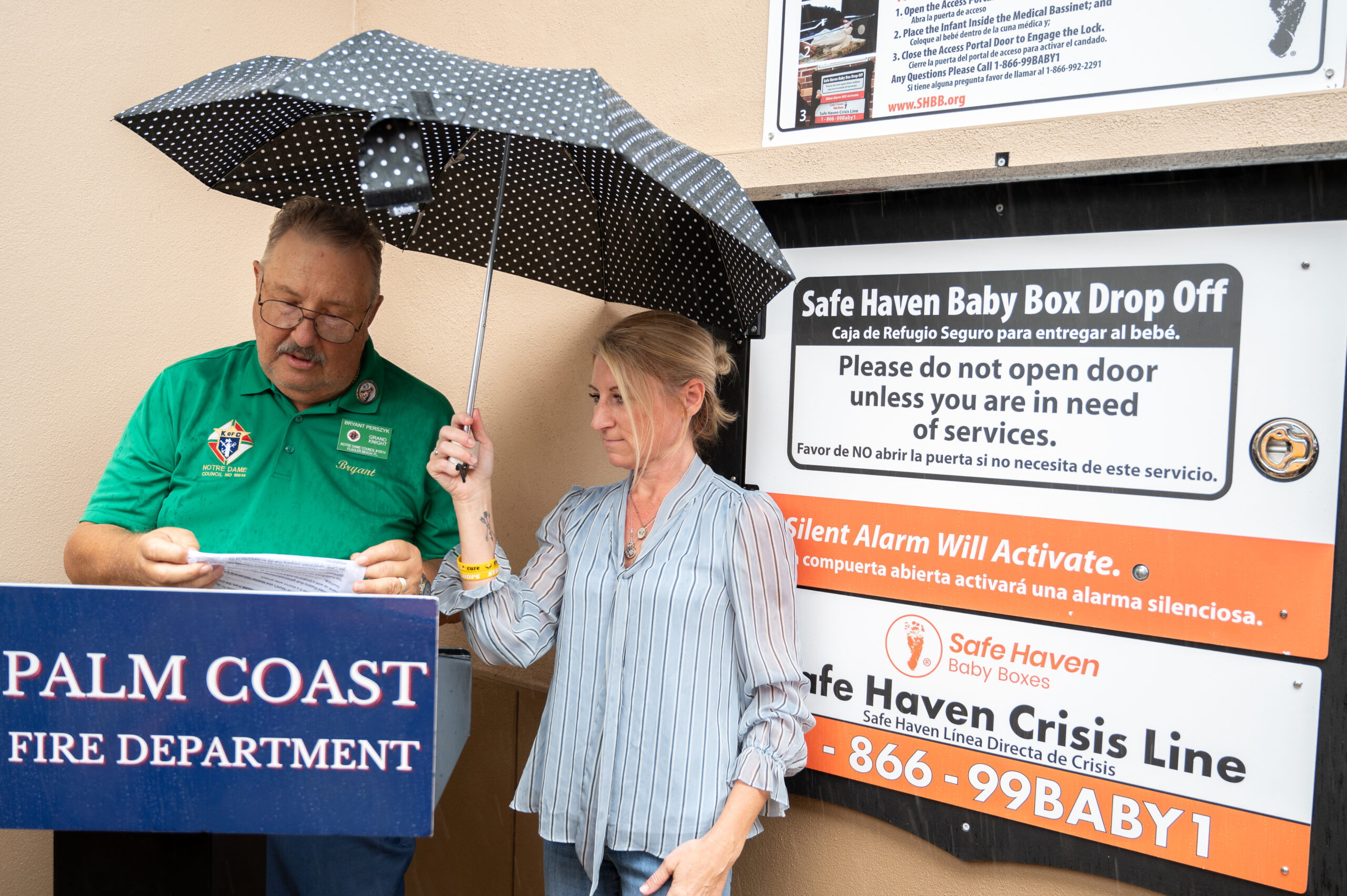 A man speaks while holding papers and a woman stands beside him holding an umbrella over them They are next to a Safe Haven Baby Box Drop Off and Palm Coast Fire Department sign on a building wall