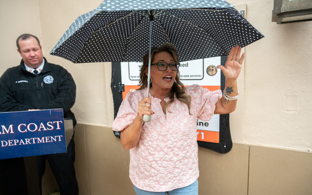 A woman in glasses and a pink blouse holds a polka dot umbrella and gestures while speaking A man in uniform stands nearby next to a podium labeled "COAST DEPARTMENT " A sign is visible in the background