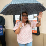 A woman in glasses and a pink blouse holds a polka dot umbrella and gestures while speaking A man in uniform stands nearby next to a podium labeled "COAST DEPARTMENT " A sign is visible in the background