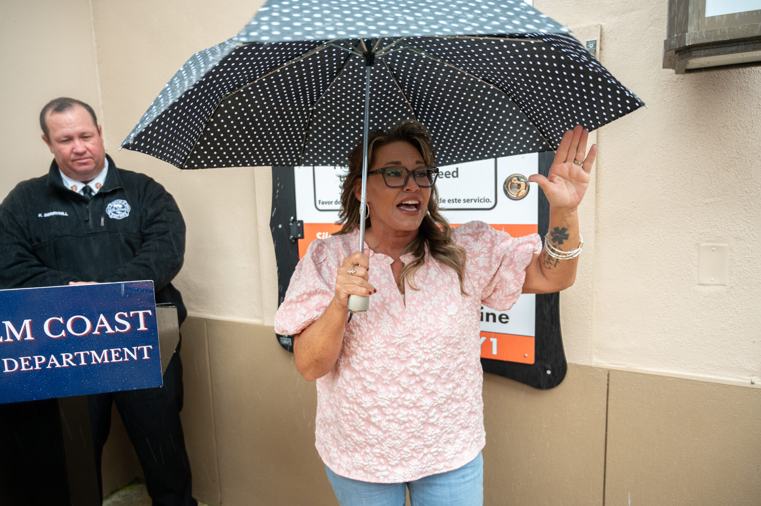A woman in glasses and a pink blouse holds a polka dot umbrella and gestures while speaking A man in uniform stands nearby next to a podium labeled "COAST DEPARTMENT " A sign is visible in the background