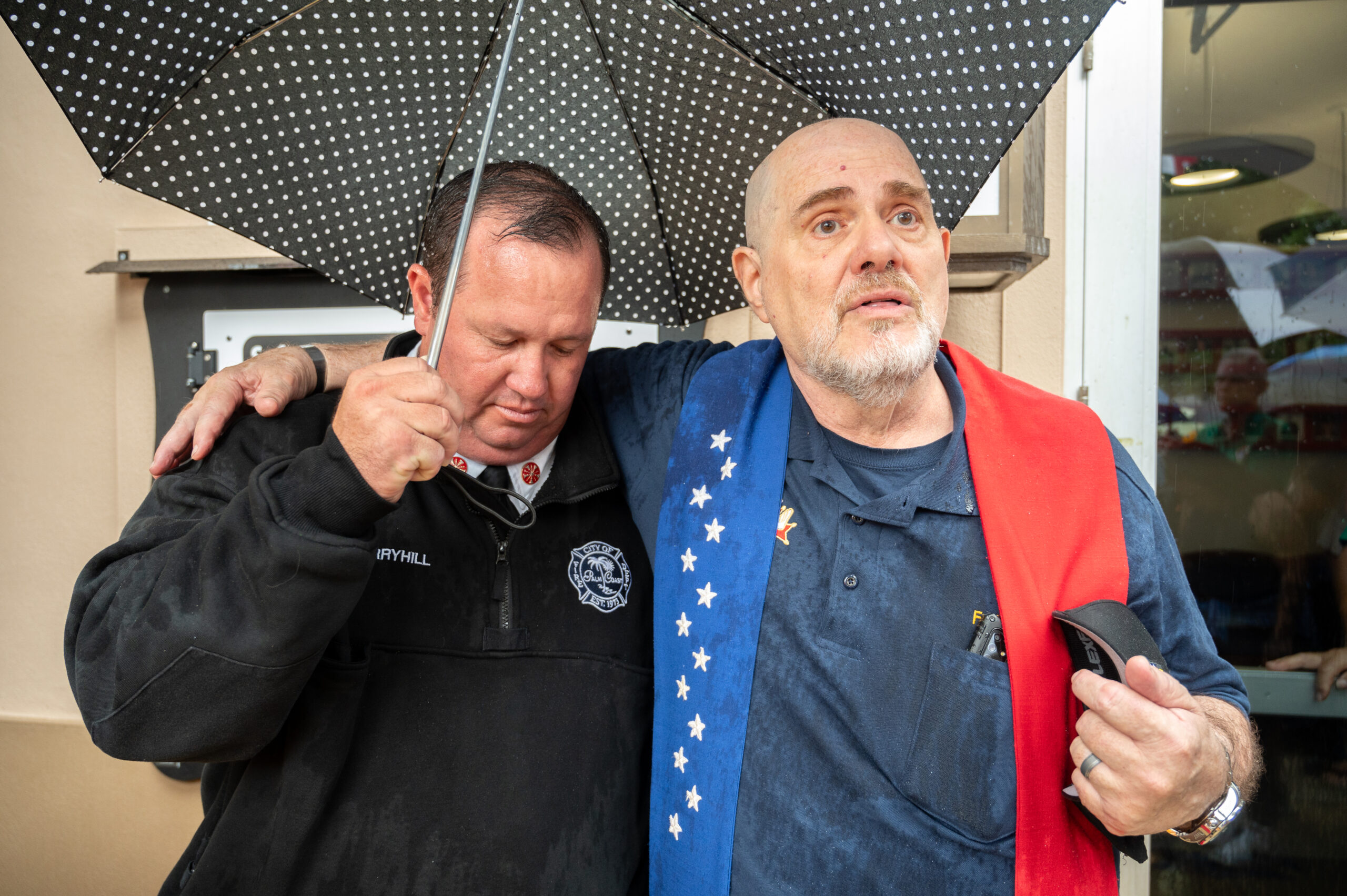Two men stand close under a black umbrella in the rain, one in a fire department uniform with his arm around the other, who looks emotional and wears a shirt draped with a red cloth featuring white stars