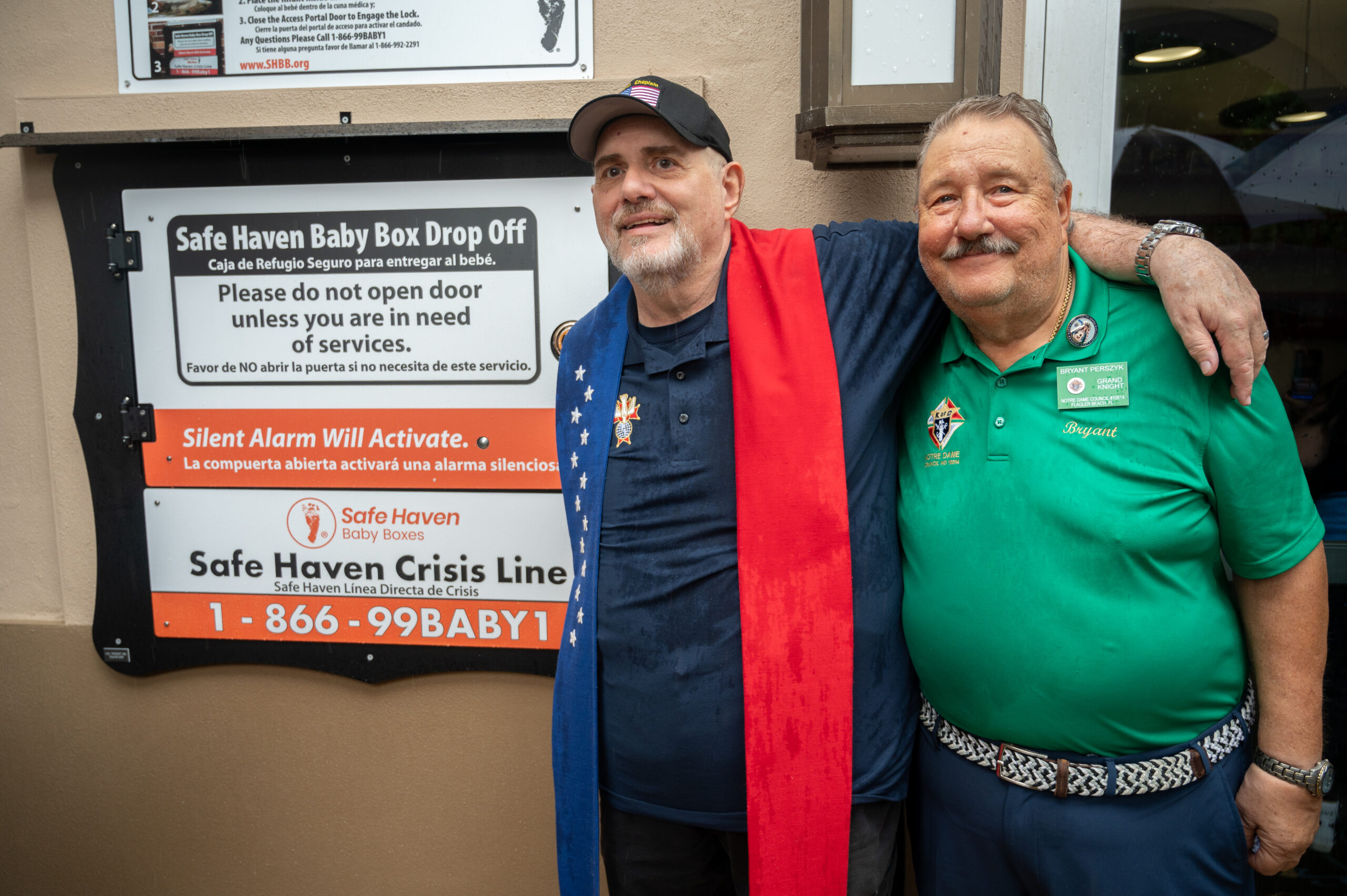Two smiling men stand arm in arm beside a Safe Haven Baby Box Drop Off sign One wears a black cap and blue jacket with a red scarf, and the other wears a green polo shirt with badges