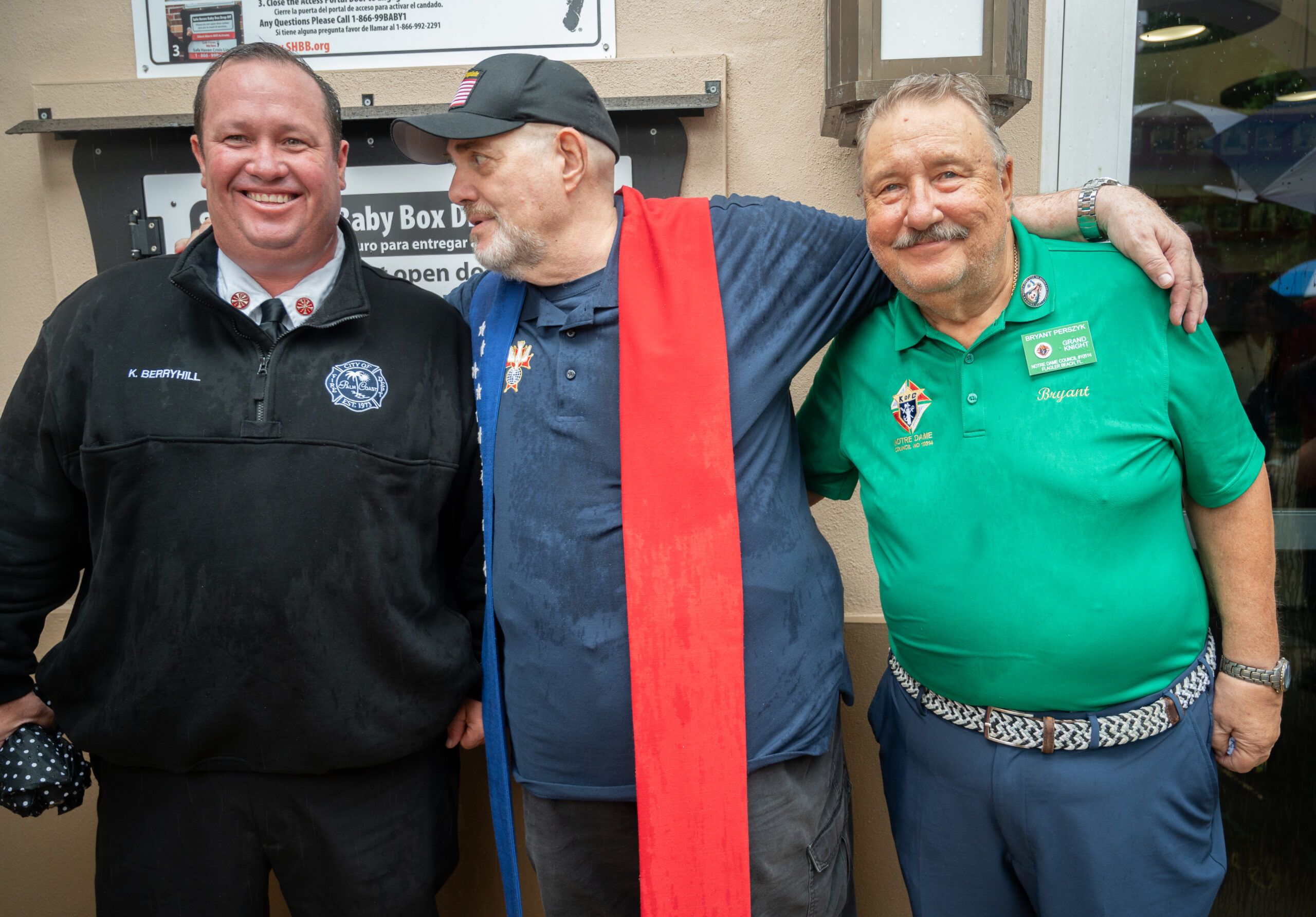 Three smiling men stand together outside One wears a black firefighter uniform, another wears a navy cap and rain soaked shirt with a red scarf, and the third wears a green polo shirt with badges They appear to be celebrating or at an event