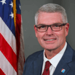 A man in a blue suit, white shirt, and red polka dot tie smiles in front of a U S flag He has short gray hair, glasses, and a pin on his lapel with a blue and red design