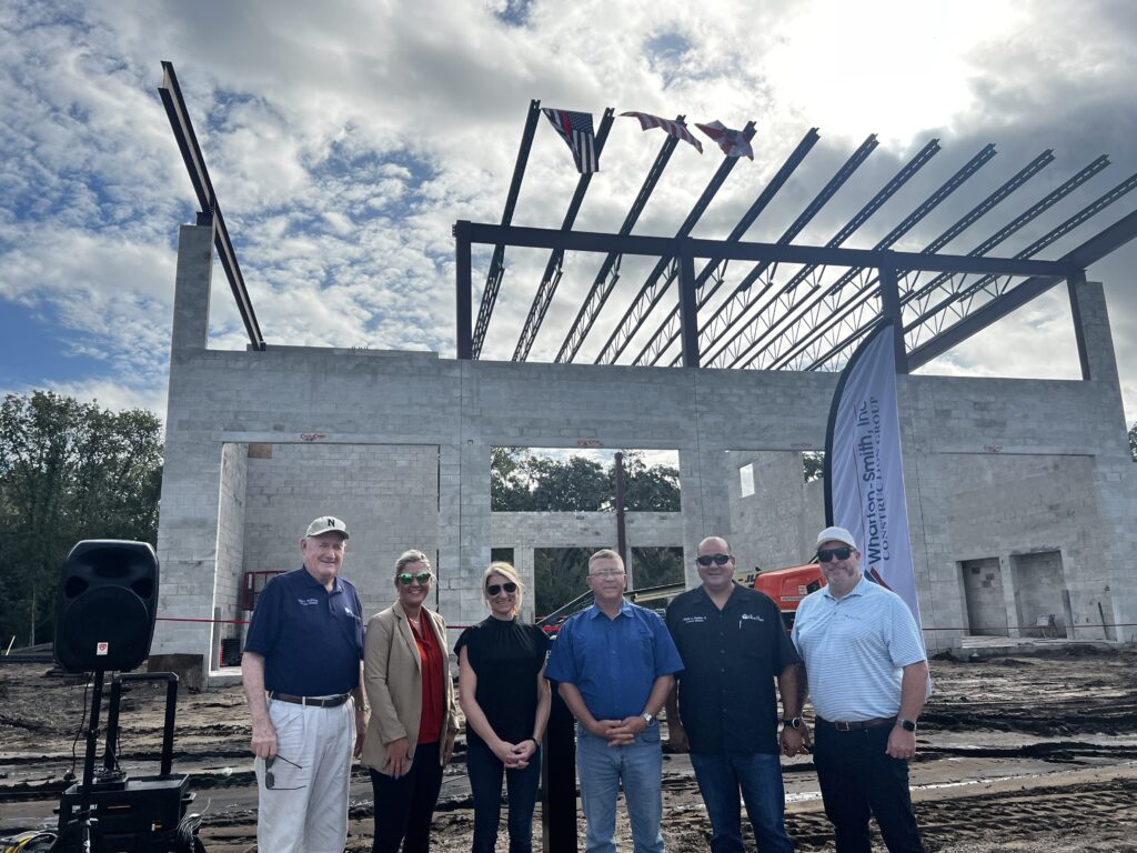 Six people stand in front of a partially constructed building with steel beams and concrete walls under a partly cloudy sky A vertical banner and construction equipment are visible in the background