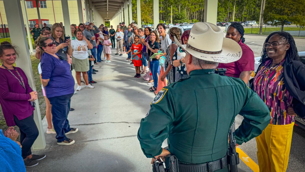 A police officer in uniform, seen from behind, stands facing a long line of smiling and engaged adults and children outside a building on a covered walkway The group appears to be participating in a welcoming or celebratory event
